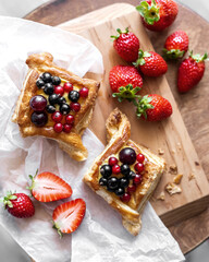 Two freshly baked pastry puffs with cream filling, berry topping and strawberry on paper and wooden table. Delicious breakfast concept. Homemade dessert. Rustic food. Top view