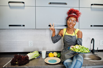 African american woman preparing healthy food at home kitchen. She hold plate with salad and show two fingers.