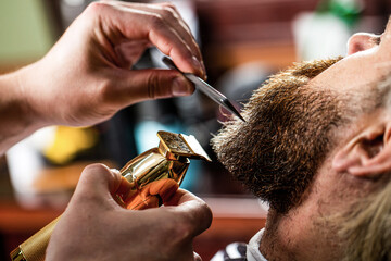 Wall Mural - Barber works with a beard clipper. Hipster client getting haircut. Hands of a hairdresser with a beard clipper, closeup