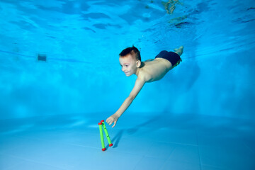 Wall Mural - Cute athletic boy dives to the bottom of the pool for a toy. Fun dives underwater. Active happy child. Swimming classes. Bodily exercises. Healthy lifestyle