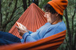 Hammock on trees in the forest ,portrait of man using mobile from inside a hammock in the forest, wears teal sweater and orange hat