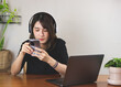 © Phuttharak - Asian woman in black shirt wearing headphones, sitting at wooden table with computer laptop, using mobile phone. white wall with house plant background.
