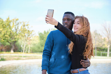 African black man and redhead caucasian woman smiling and taking a selfie with their smartphone in a park. Young multiracial couple portrait.