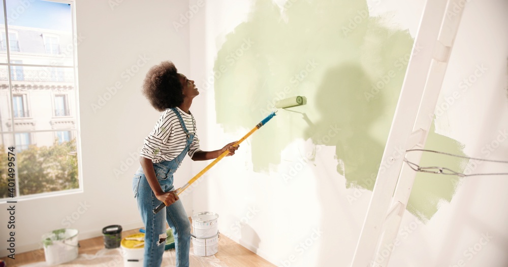 Young African American woman dancing and painting wall with roller brush while renovating apartment. Rear of female having fun redecorating home, renovating and improving Repair and decorating concept - obrazy, fototapety, plakaty 