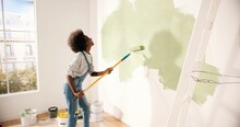 Young African American Woman Dancing And Painting Wall With Roller Brush While Renovating Apartment. Rear Of Female Having Fun Redecorating Home, Renovating And Improving Repair And Decorating Concept