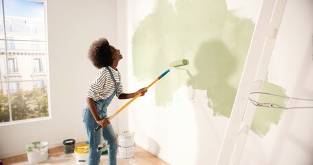 young african american woman dancing and painting wall with roller brush while renovating apartment.