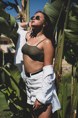 Wall Mural - cute young teenage girl or woman posing with banana tree leaves dressed in summer hipster vintage white stylish clothes. Trendy girl posing. Funny and positive woman in sunglasses