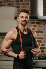 Wall Mural - A muscular man in a tank top with a pull elastic rope on the shoulders is posing in his apartment. The bodybuilder with tattoos on his forearms is demonstrating his sporty physique at home