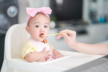 Asian cute baby girl in a pink bib and hair bow sitting on a white high chair. Her mother giving healthy food by spoon in the morning at home