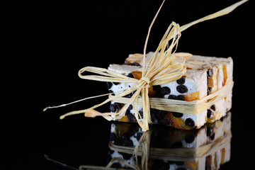 Several pieces of torrone or white nougat with nuts, candied fruits and chocolate are tied together with raffia and lie on a reflective surface in front of a dark background