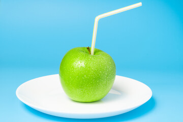 Green fresh apple with water drops. Juicy apple with straw on the plate on cheerful blue background. Natural apple juice concept.