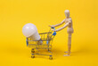 Wooden puppet rolls shopping cart with led light bulb on yellow background