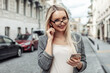 © splitov27 - Portrait of confident business woman in urban street. Blond girl in business suit using smartphone and listening to music with wireless headphones