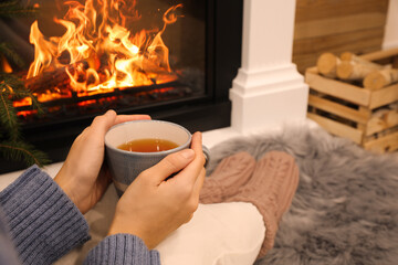 Wall Mural - Woman with cup of tea sitting near burning fireplace, closeup