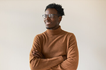 Wall Mural - Studio shot of handsome enthusiastic dark skinned guy in trendy eyeglasses having inspired facial expression, thinking about future, making plans, keeping arms crossed on his chest and smiling