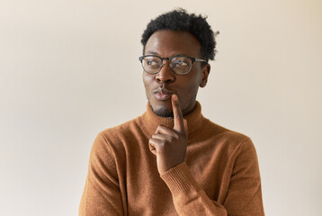 Wall Mural - Studio image of handsome stylish young black man in trendy glasses holding finger at lips and looking away with mysterious smile, thinking big, brainstorming, having brilliant idea in his mind