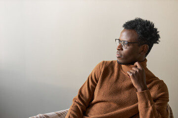 Wall Mural - Isolated shot of fashionable young African American man in elegant turtleneck sweater and spectacles sitting on chair against copy space black wall background, looking away with thoughtful expression