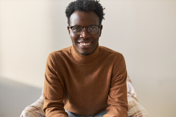 Wall Mural - Positive energetic young dark skinned student smiling, happy after passing exams with excellent marks, siting in comfortable armchair against blank wall background with copy space for your content