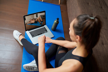 Wall Mural - A laptop screen view over a woman's shoulder. A fit girl in a tight suit is watching a workout video on a laptop in her apartment. Woman is listening to a coach during an online fitness class at home