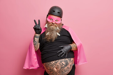 portrait of funny bearded superhero makes peace gesture has big tattooed belly wears mask helmet and