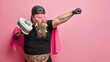 © wayhome.studio  - Studio shot of self confident plump man superhero stretches arm holds plunger wrapped with toilet paper has tattooed belly wears costume poses against pink background. Copy space for promotion