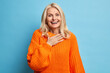 Portrait of sincere blonde woman smiles broadly has white perfect teeth keeps hand on chest feels very glad to receive help from close person wears orange knitted sweater isolated over blue background