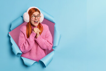 Wall Mural - Pretty cheerful ginger girl keeps hands together and looks with happy impressed expression aside dressed in warm winter sweater and earmuffs concentrated aside on awesome advertisement poses in hole
