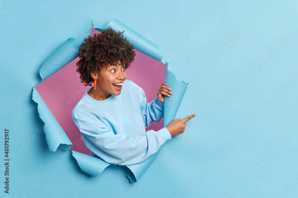 Cheerful dark skinned woman with curly hair dressed in casual sweater indicates at copy space shows amazing offer breaks through paper background expresses positive emotions. Your advertisement there - obrazy, fototapety, plakaty 