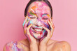 Portrait of brunette young Asian woman smiles pleasantly keeps both hands on cheeks stands with closed eyes has face smeared by colorful oil paints isolated over rosy background. Creative makeup
