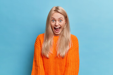 Wall Mural - Portrait of stunned blonde woman reacts on awesome news keeps mouth widely opened looks in stupor cannot believe her eyes wears orange knitted sweater isolated over blue background. Omg concept