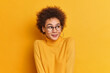 Lovely curly haired girl looks aside with glad expression wears casual turtleneck transprent glasses admires something beautiful feels happy isolated over yellow background. Emotions concept