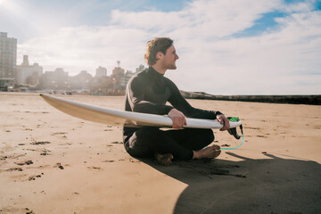 Surfer sitting on sandy beach with surfboard.