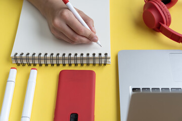 Wall Mural - A high angle shot of a person taking notes in a notebook with headphones, pens and a laptop on a yellow surface