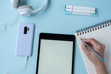 Wall Mural - A high-angle shot of a person taking notes with a tablet, mobile phone, headphones, and markers on a blue desk