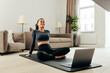 © Artem Varnitsin - Smiling woman in sport clothes sitting on a mat in living room in front of a laptop. Young female relaxing during training at home.