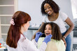 © sofiko14 - Dental treatment without fear. Female dentist in whilte uniform and gloves, examining teeth of little African American girl, while her mother supporting her behind, at modern pediatric dental clinic