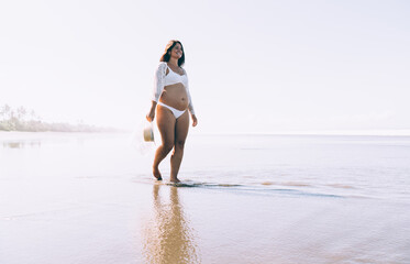 Smiling young pregnant lady in bikini walking on sandy beach