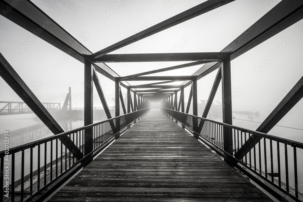 A grayscale shot of light at the end of a modern beautifully architectured bridge surrounded by fog - obrazy, fototapety, plakaty 