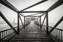 A Grayscale Shot Of Light At The End Of A Modern Beautifully Architectured Bridge Surrounded By Fog