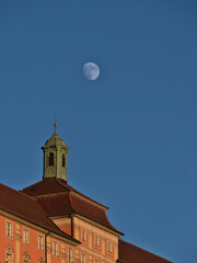 Old building with pink colored decorated facade and bell tower on the roof in beautiful evening sun with moon visible in the blue and clear sky in town Meersburg, Lake Constance, Germany.