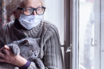 Elderly woman in protective mask holding a cat looks out the window wearing. Christmas quarantine covid19. Coronavirus epidemic. Waiting. Depression. Insulation at home. Pets save you from loneliness