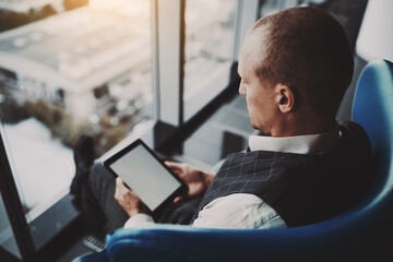 Sideview of a successful man entrepreneur with a digital tablet in hands with empty screen mock-up, sitting on an armchair of a classic blue color on the top floor of a luxury business skyscraper