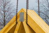 Fototapeta Tęcza - Part of a wooden house roof section, waiting to bi raised and assembled. Corner binding made of metal being raised by a crane.