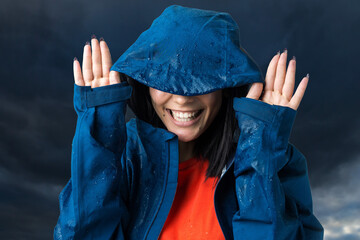 Portrait of a smiling girl dressed in blue raincoat in drops posing with hood against the background of the dramatic sky. Bad weather concept.