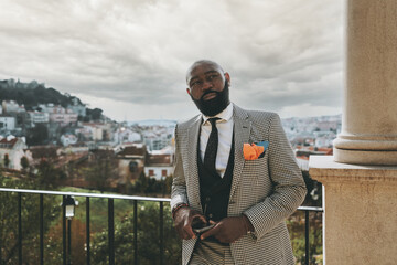 A portrait of a stately mature bearded black guy in a plaid elegant costume with a necktie, he is leaning against a metal fence on the street, on high ground, with a cityscape in the background