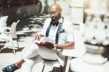 A Portrait Of A Fancy Mature Bald Bearded Black Man In Eyeglasses And A White Elegant Suit With A Bow-tie, Reading A Book With Multiple Bookmarks While Sitting In An Outdoor Restaurant On A Sunny Day