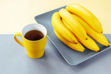 yellow cup of tea and bananas on gray plate. trendy colors 2021.