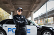 confident policewoman with crossed arms looking at camera near patrol car on blurred background on urban street.