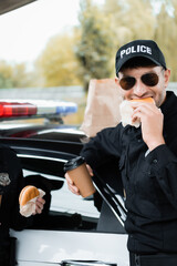 Police officer eating burger and holding coffee to go near colleague and car on blurred background.