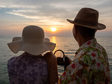 Rear View Of An Old Couple With The Hats Looking Out To Orange Ocean Sunset.